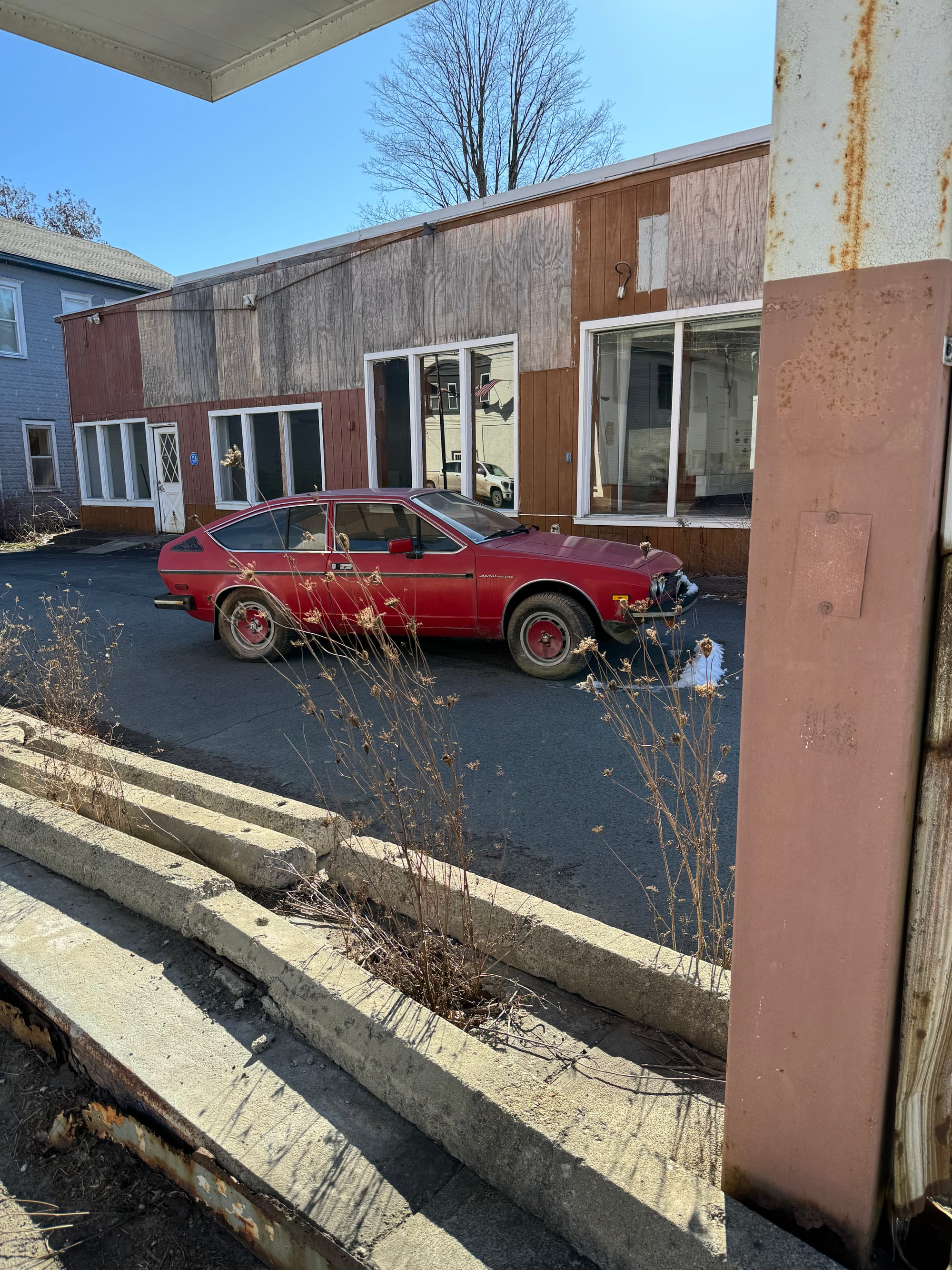 A red fastback car from the 1980s, sitting semi-abandoned in front of an abandoned building in Roscoe, New York.